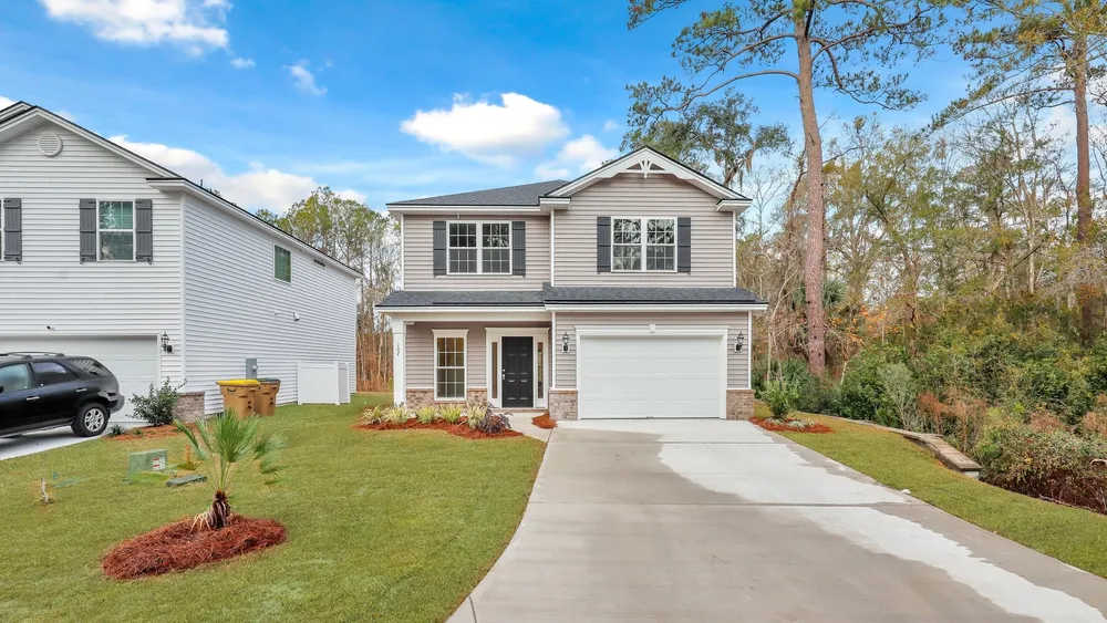 Front view of the Hatteras C with taupe siding, dark shutters, a decorative front gable, a covered entry porch, and an attached garage beside a landscaped lawn and wooded backdrop.