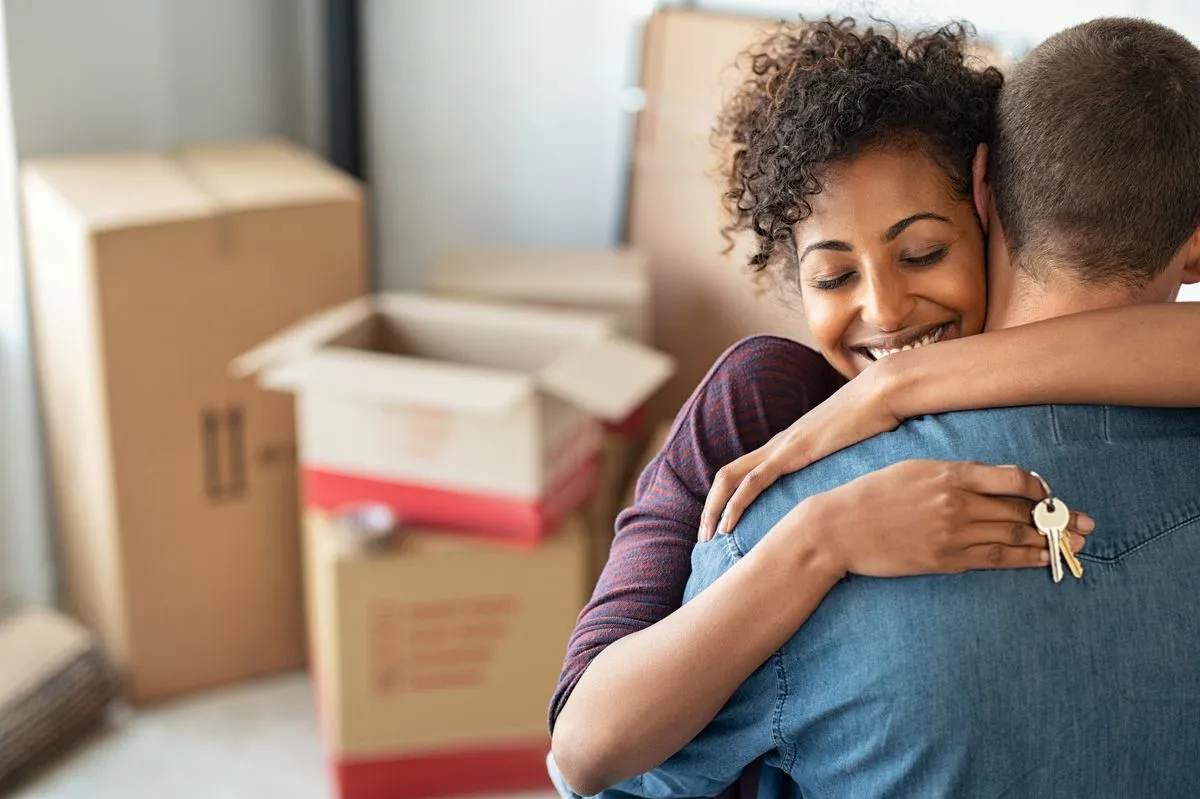 Two people hugging while holding keys with moving boxes in the background