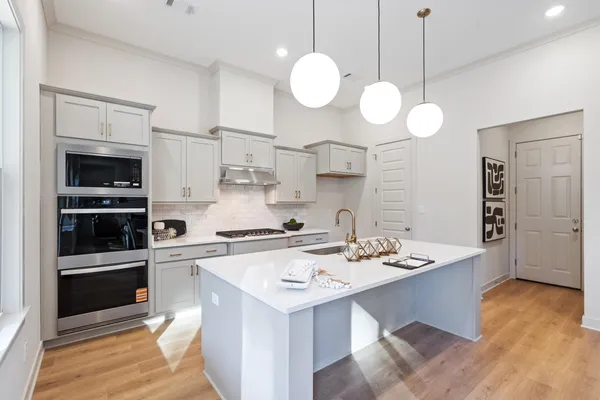 Kitchen featuring gray cabinetry, stone countertops, a center island with a built-in sink, stainless steel double wall ovens, and globe pendant lighting