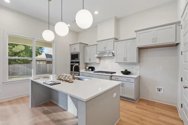 Kitchen featuring an oversized central island with stone-surface countertop, light grey cabinetry, subway-style backsplash, integrated wall oven, and pendant globe lighting