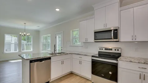 A sleek kitchen with stainless steel appliances and light-colored cabinets and countertops, overlooking an open-concept living space and café area in the newly constructed home by Ivey Homes at 854 Rachel Branch Forrest Bluff, North Augusta, SC 29841.