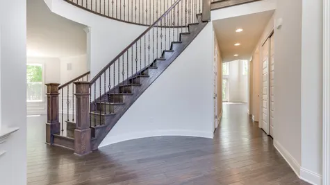 Curved stair case in front hall of The Stoneleigh luxury home model, with dark hardwood floors & stairs, decorative handrail.