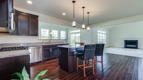 The Wexford Kitchen with dark cabinets, island, wood floors, and carpeted family room with optional fireplace beyond.