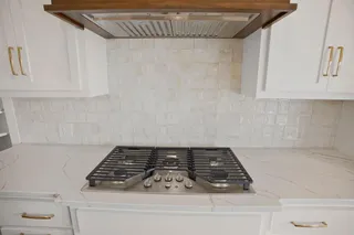 White cabinetry with gold-finish hardware, built-in gas cooktop, patterned tile backsplash, and a wood-finish range hood